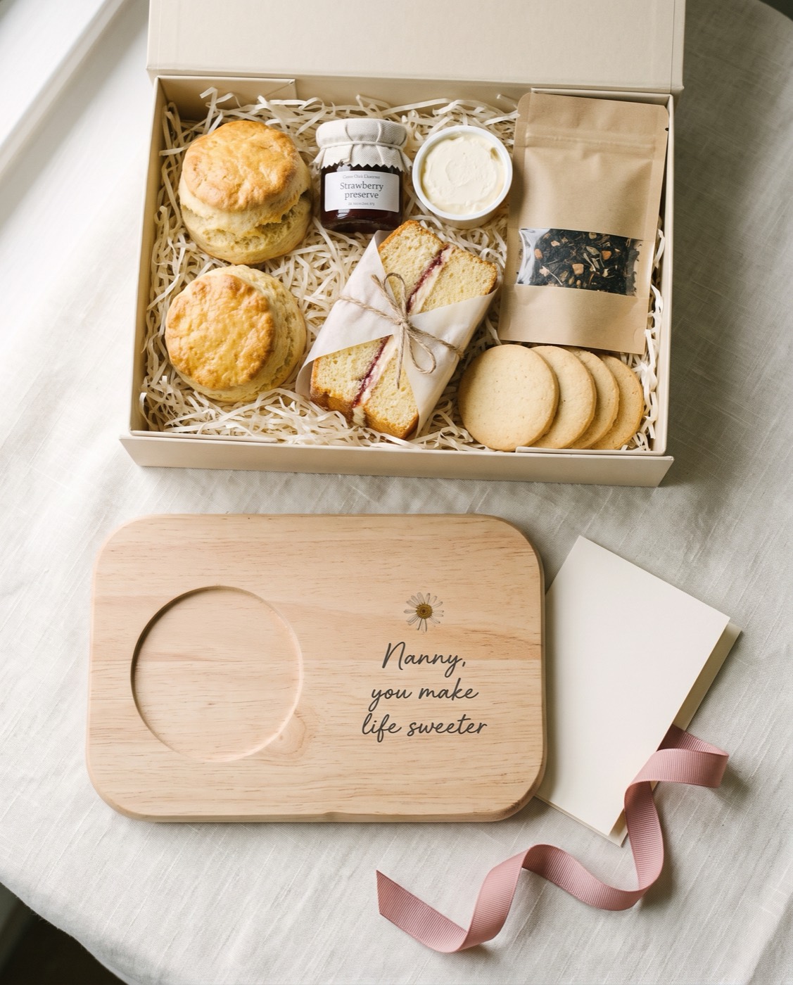 An open gift box of scones, jam, clotted cream, cake, tea and biscuits, with a beech board printed with the words Nanny, you make life sweeter